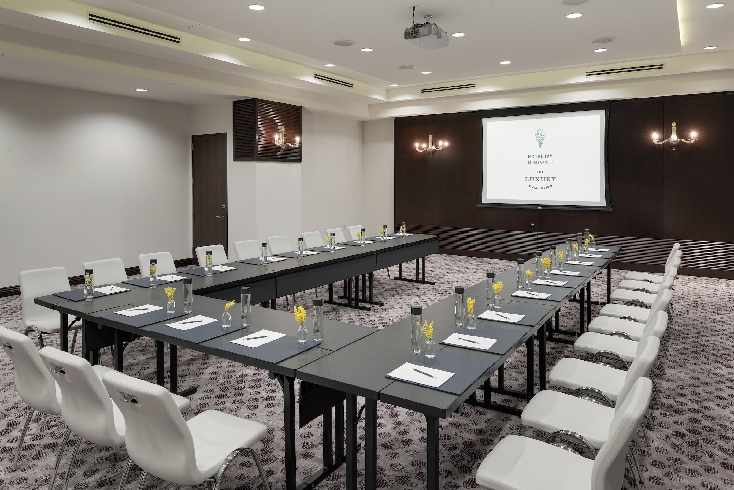 An elegantly designed meeting room at Hotel Ivy’s Grand Salon, set up in a U-shape conference style with white chairs, dark tables, and a presentation screen on wooden coloured wall