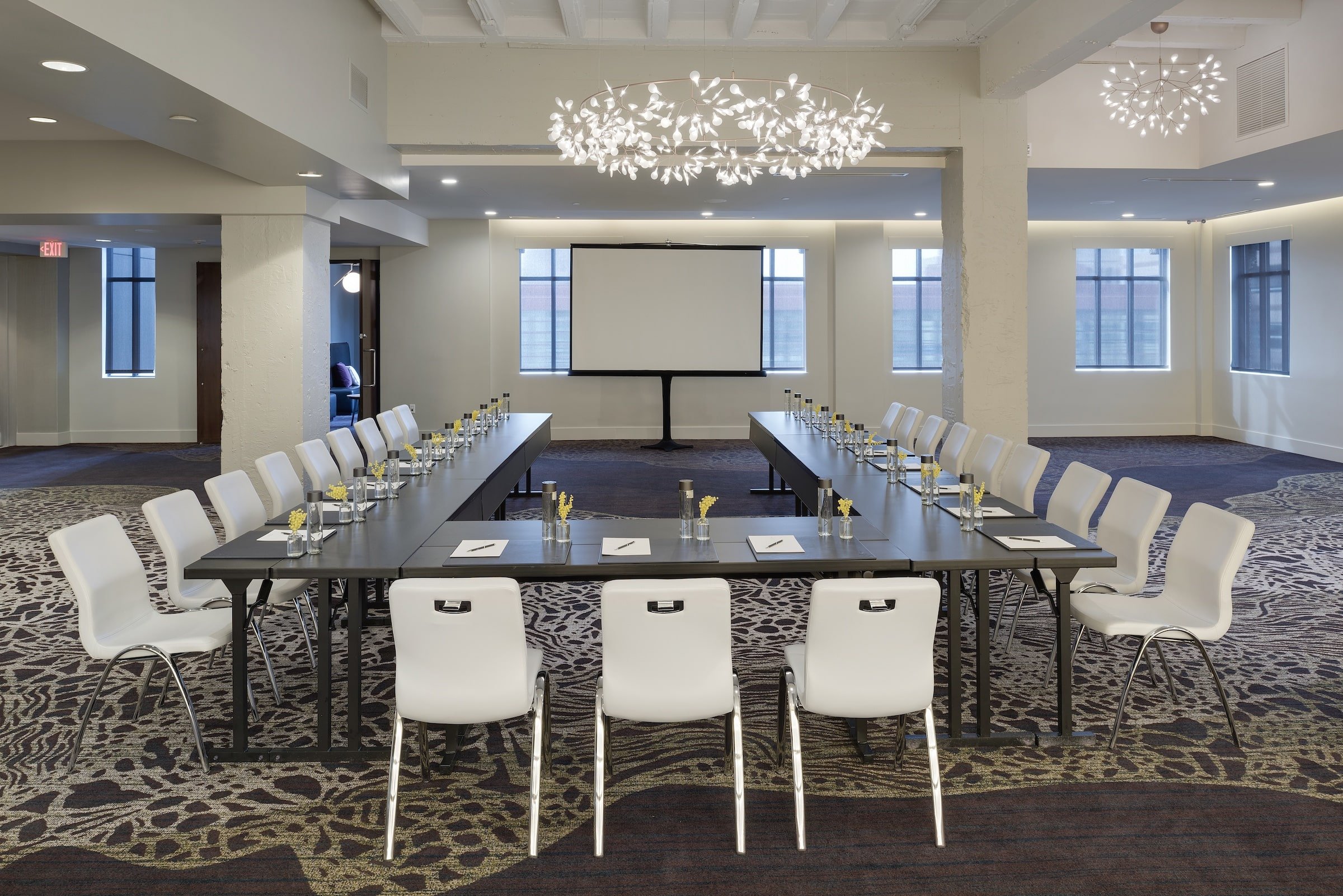 A spacious and elegantly designed meeting room at Hotel Ivy’s Grand Studio, set up in a U-shape conference style with white chairs, dark tables, and a presentation screen.
