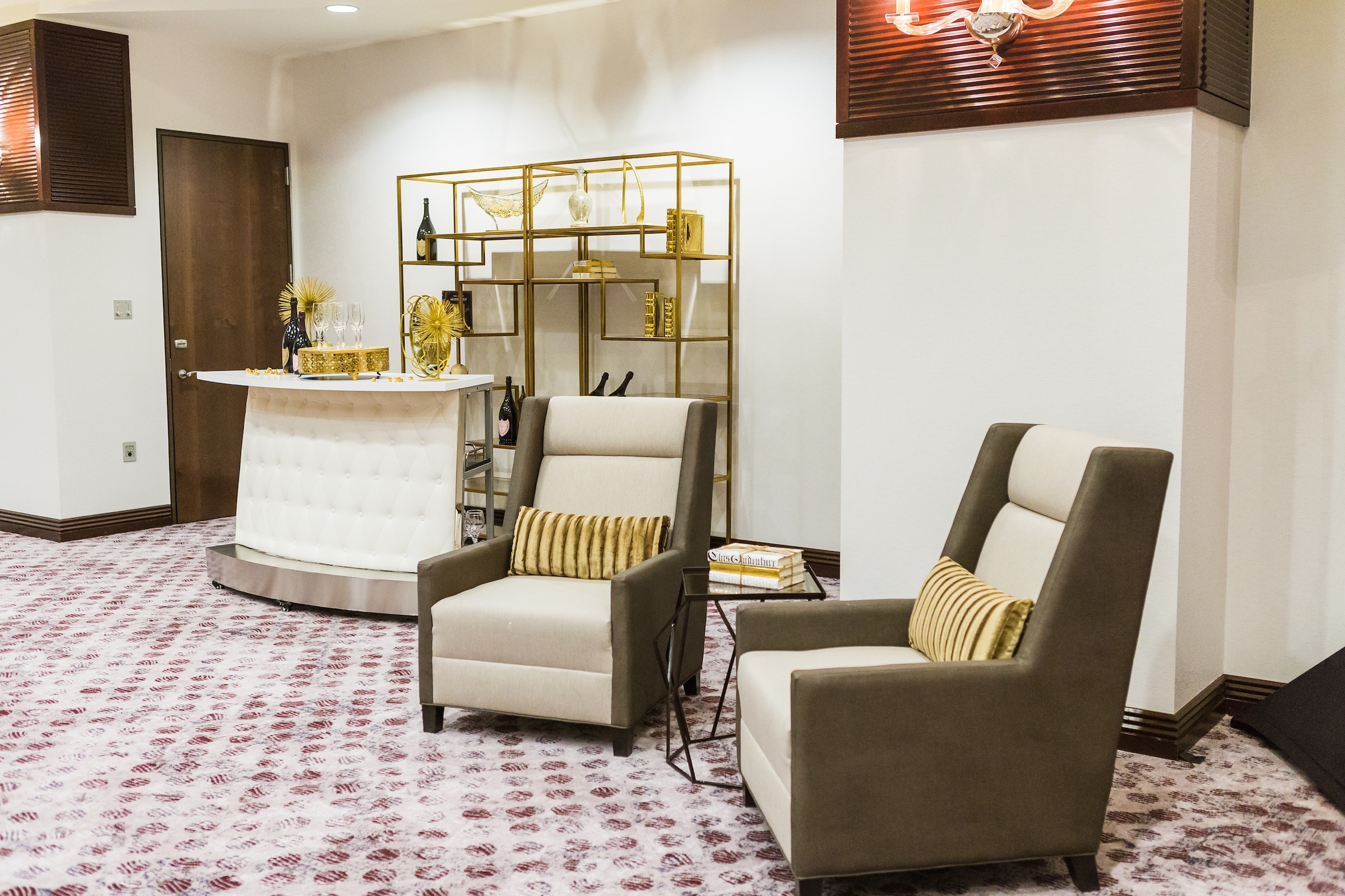 Liquor tables with golden decorations on a white reception table, two comfortable couches and a glass tea table with books in Pre-function Space at Hotel Ivy