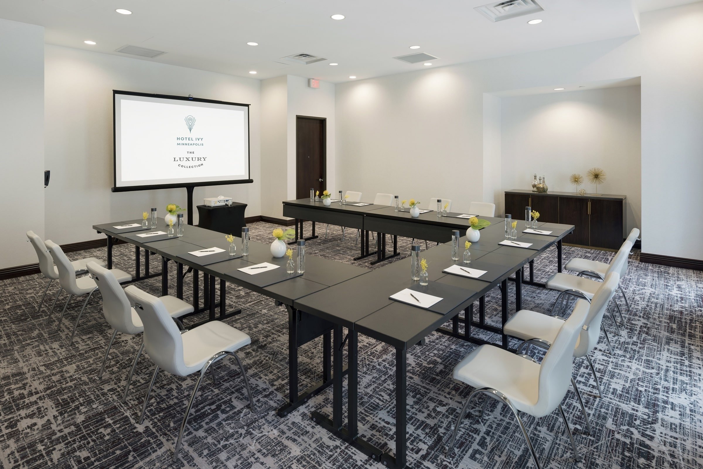 An elegantly designed meeting room at Hotel Ivy’s Grand Salon, set up in a U-shape conference style with white chairs, dark tables, and a presentation screen on wooden coloured wall