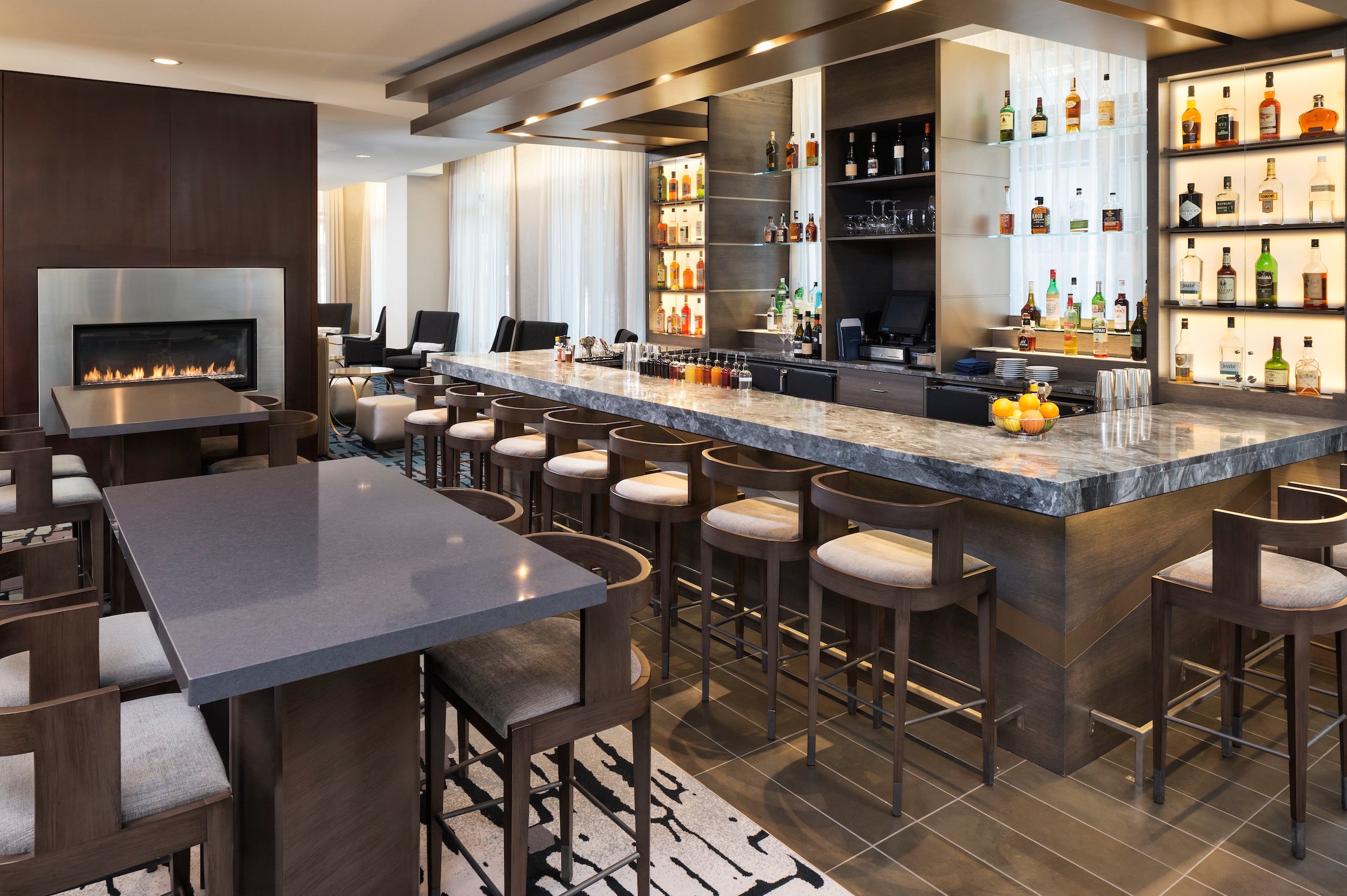 U-shaped of bar table with liquor selections and high tables with decorations of fireplace on dark brown wall at Bar Edera of Hotel Ivy