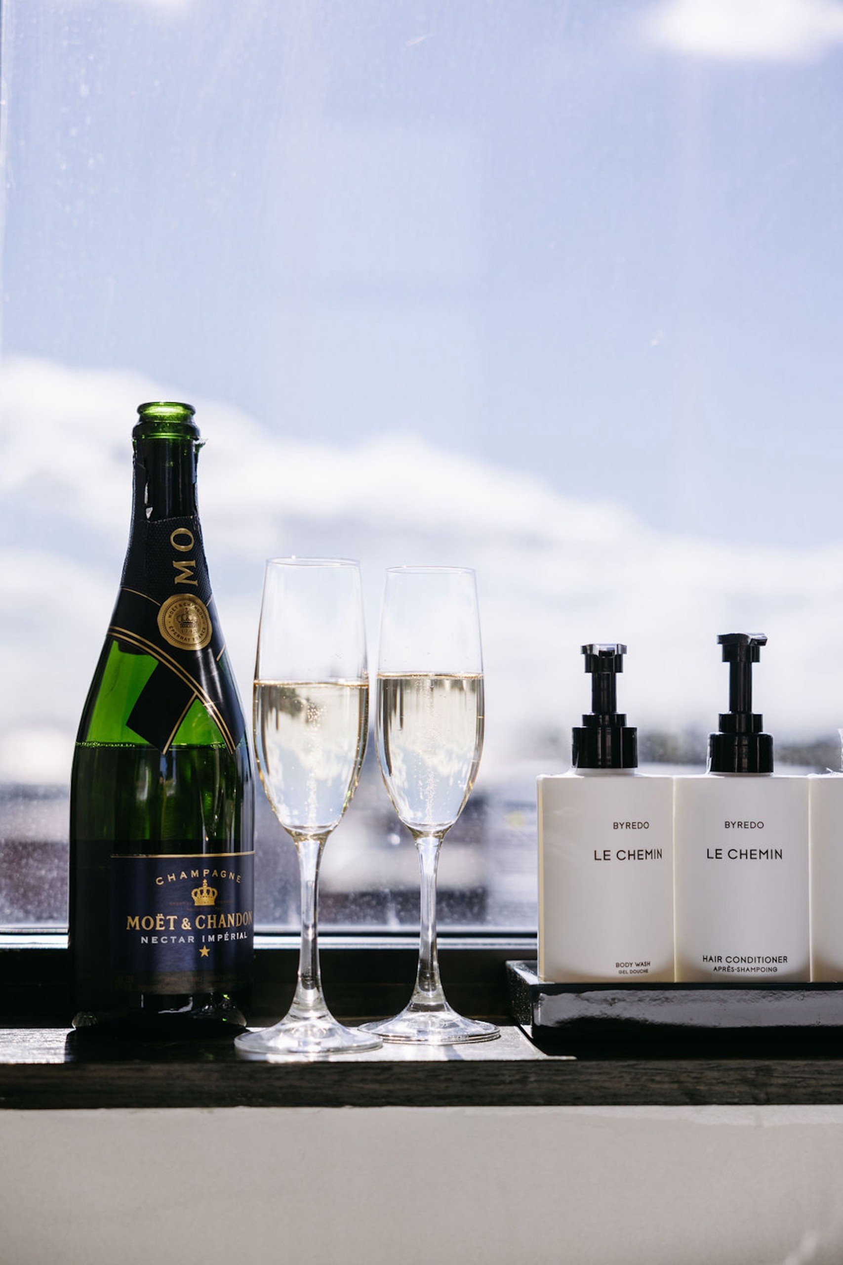 A Champagne bottle, two glasses filled with champagne, and amenities on a large window having sunshine at Hotel Ivy